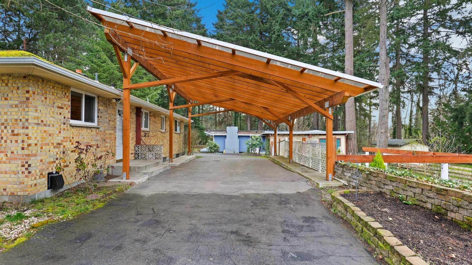 Wooden pergola covers driveway beside brick house surrounded by tall trees