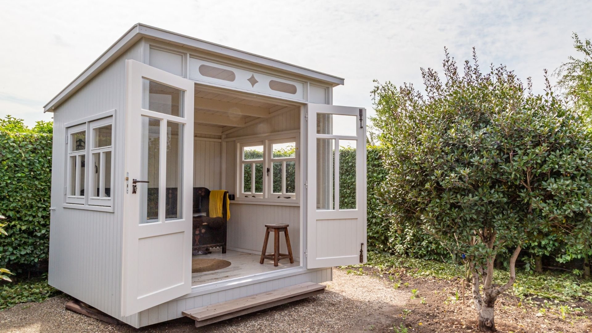 Modern white garden studio with open glass doors, stool, and surrounding hedges