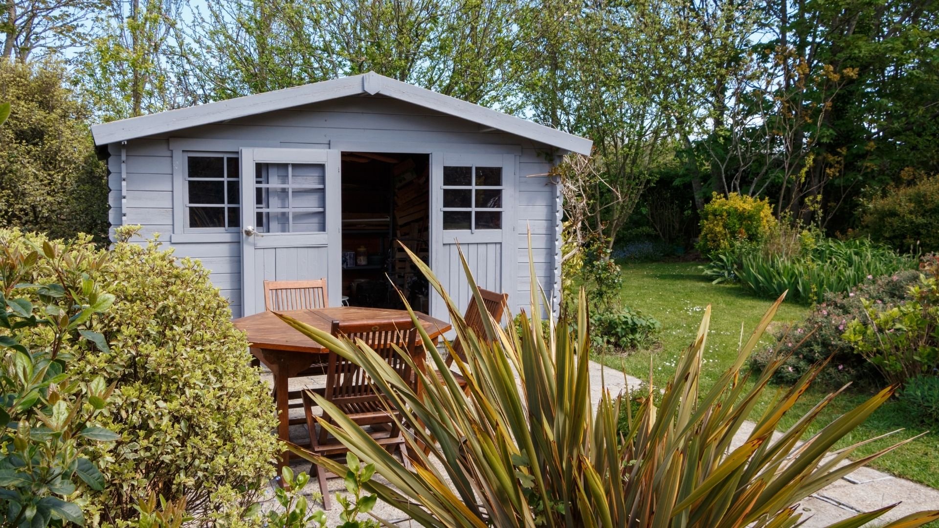 Light blue garden shed with wooden deck, tables, and surrounding greenery and plants.
