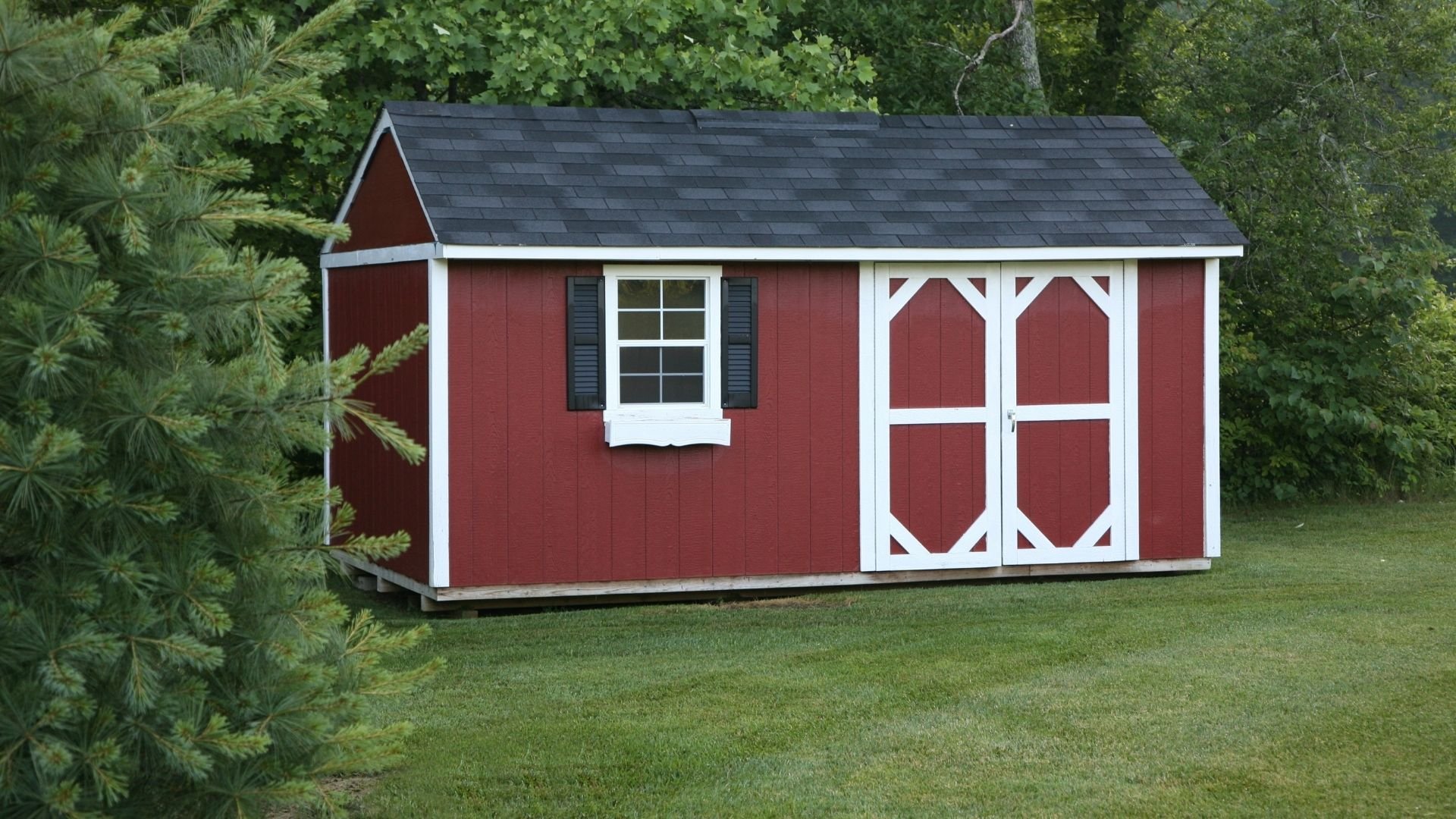 Red barn shed with white trim, dark roof, window, and double doors in grassy yard