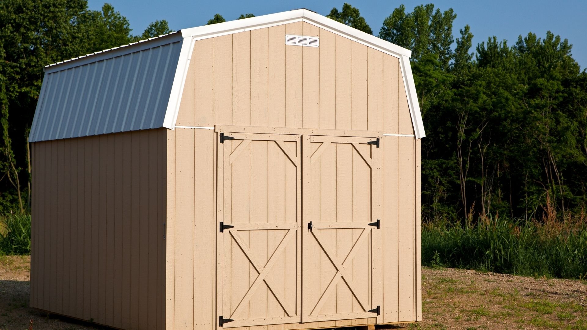 Beige wooden barn shed with gambrel roof and double doors surrounded by trees.