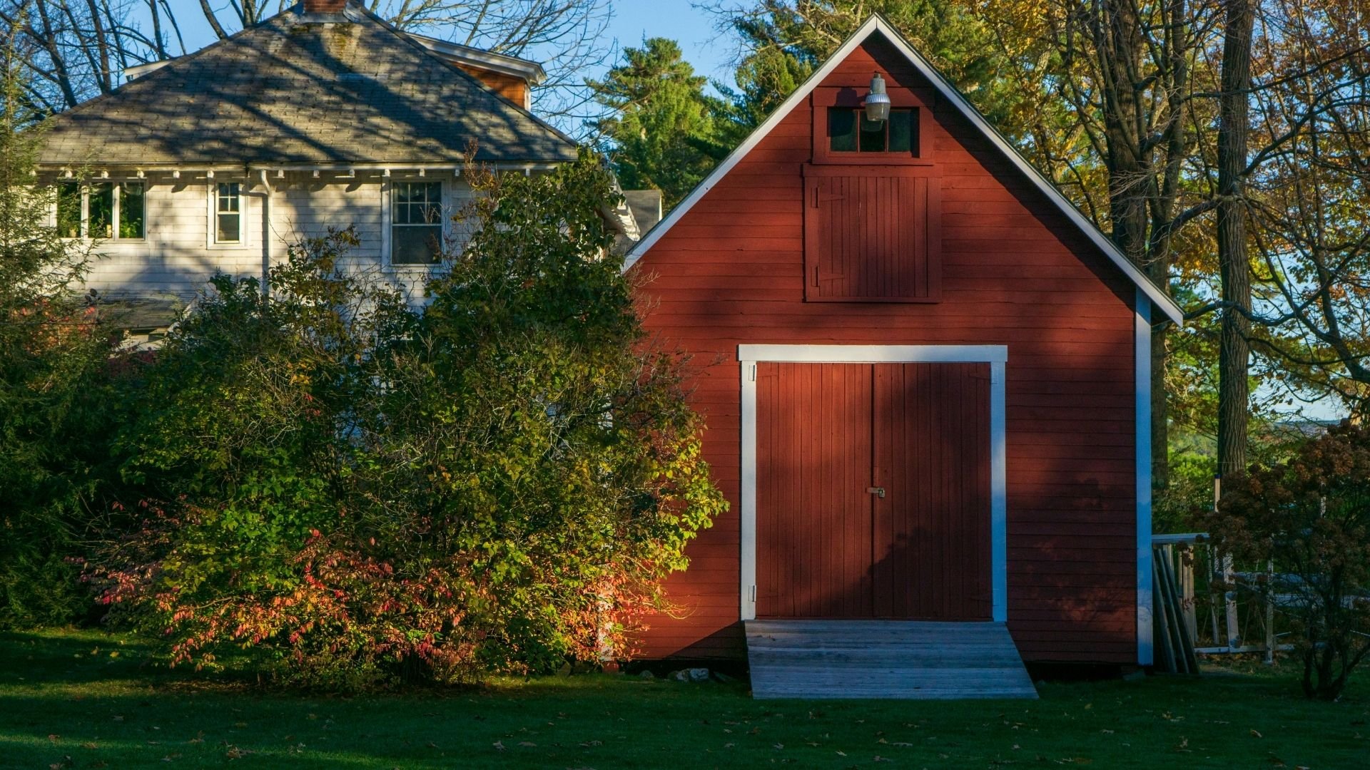 Red barn and white cottage surrounded by autumn foliage and trees under blue sky