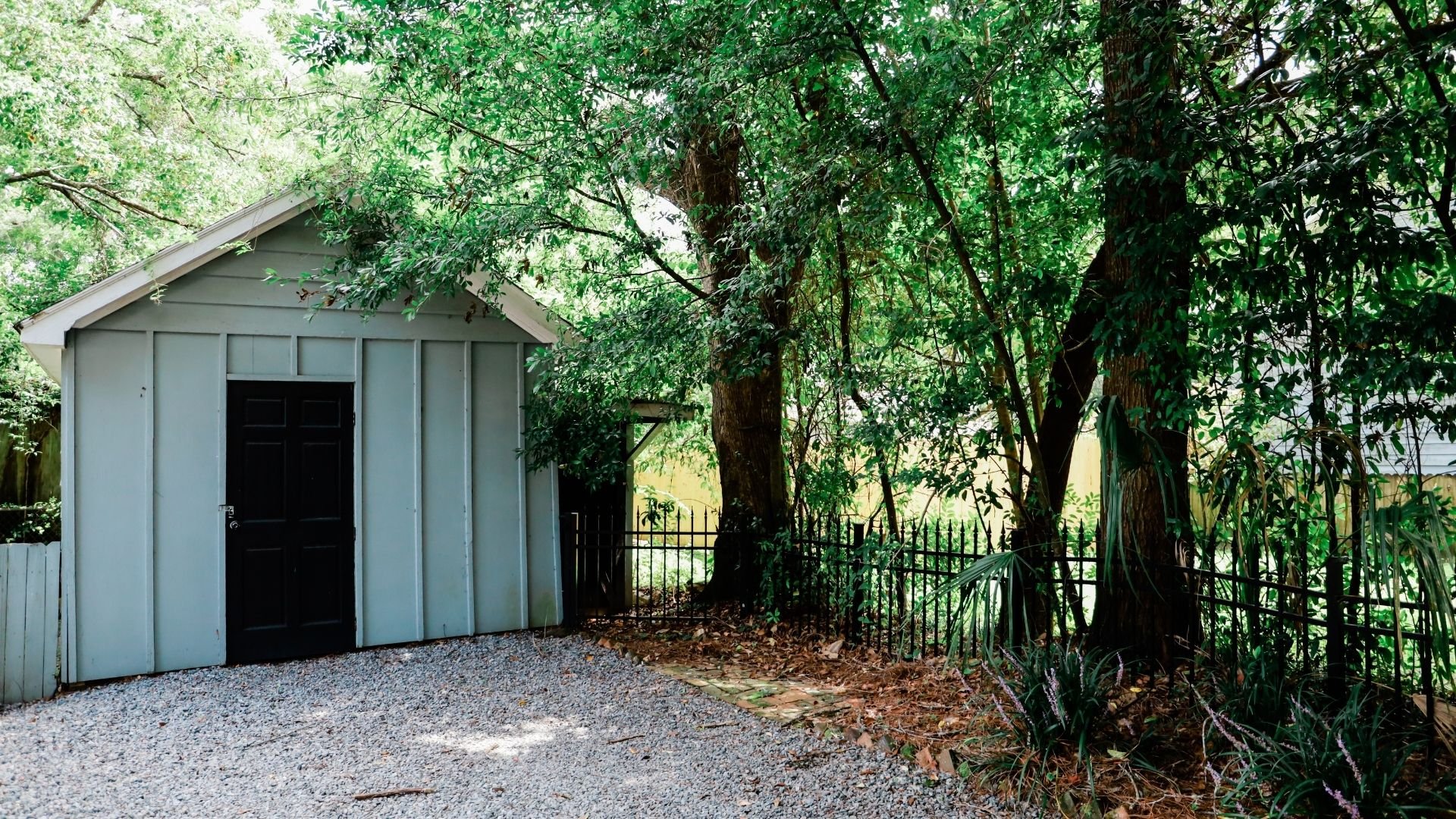 A gray storage shed nestled beneath tall trees with dense green foliage and iron gate
