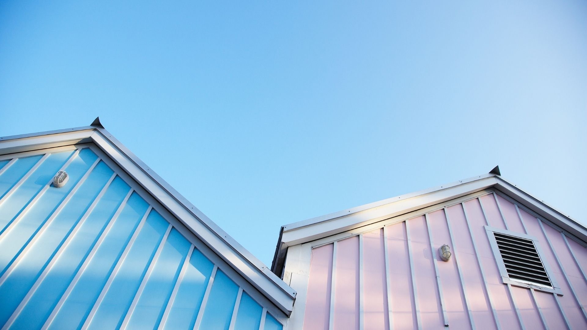 Two colorful beach huts with blue and pink corrugated metal roofs against clear sky.