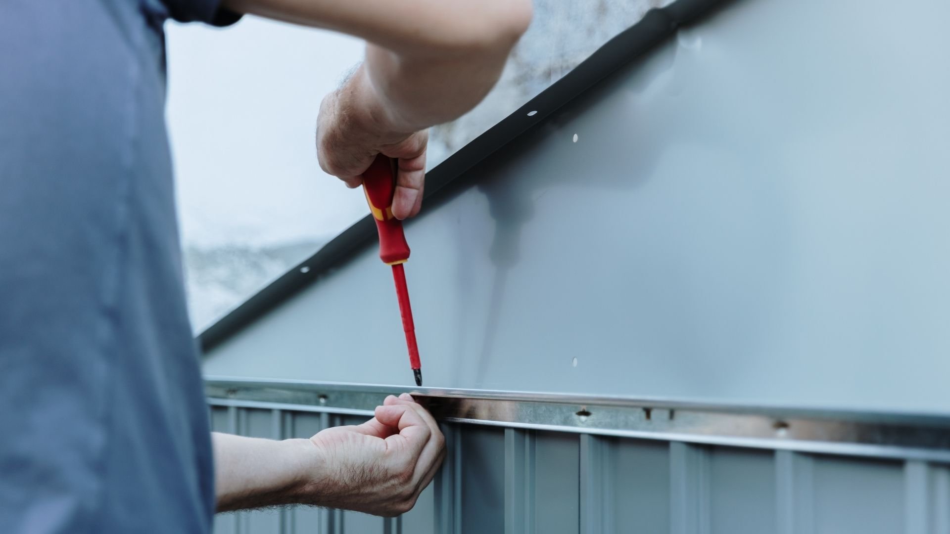 Person holding red screwdriver, performing maintenance on metal railing outdoors.