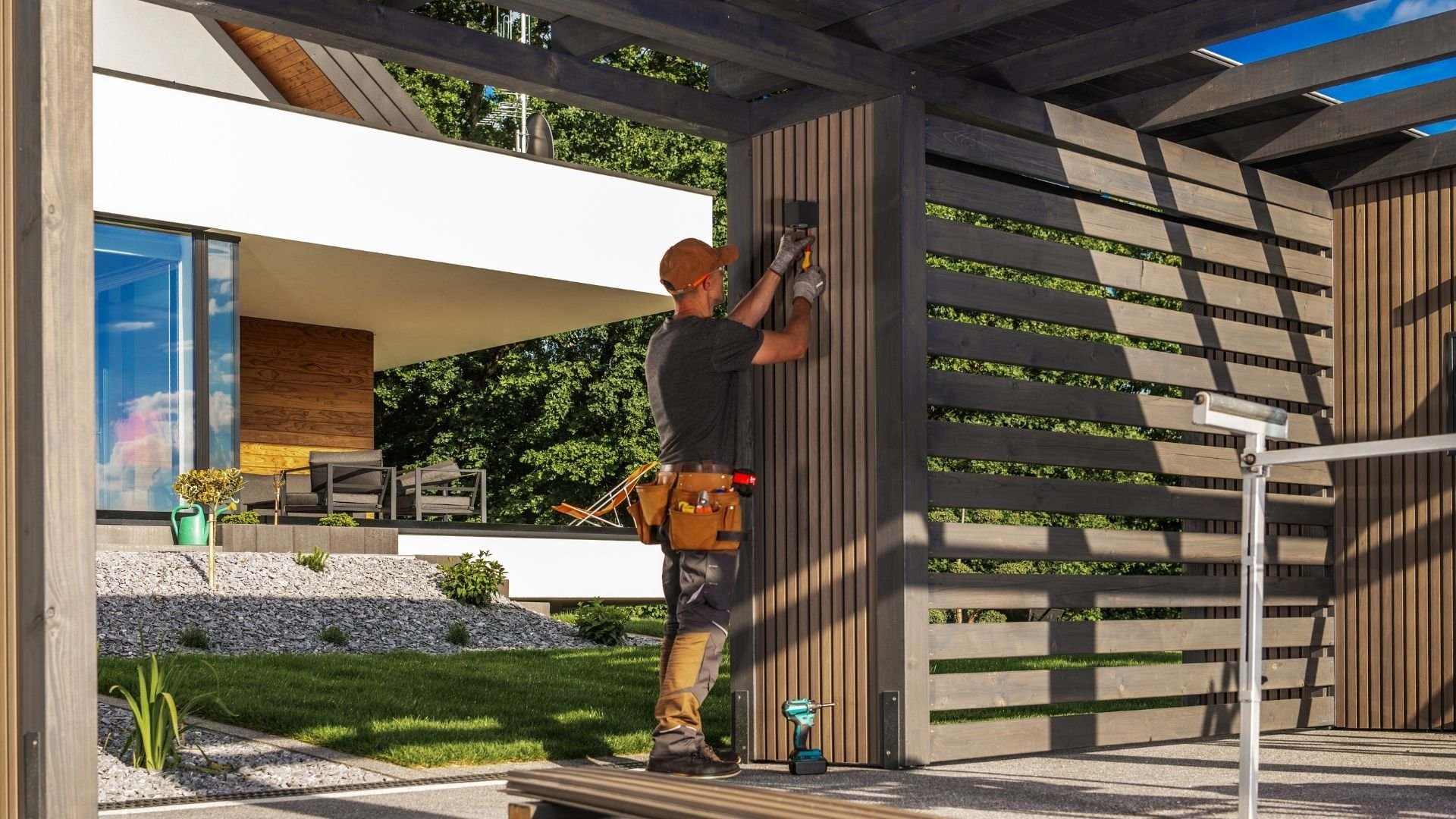 Worker installing vertical wooden slats on pergola structure near modern house exterior
